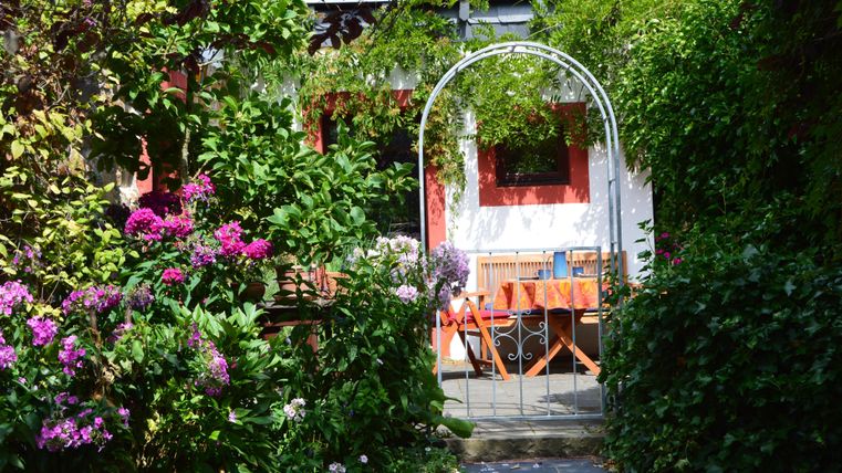 Ein schöner Garten mit bunten Blumen und einem begrünten Tor. Im Hintergrund steht ein Tisch mit Stühlen, bereit für gemütliche Stunden.