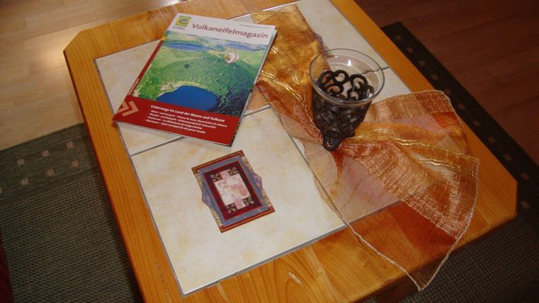 Ein Holztisch mit einer bunten Tischdecke. Darauf liegen ein Reiseführer, eine kleine dekorative Box und ein Glas mit Deko-Elementen.