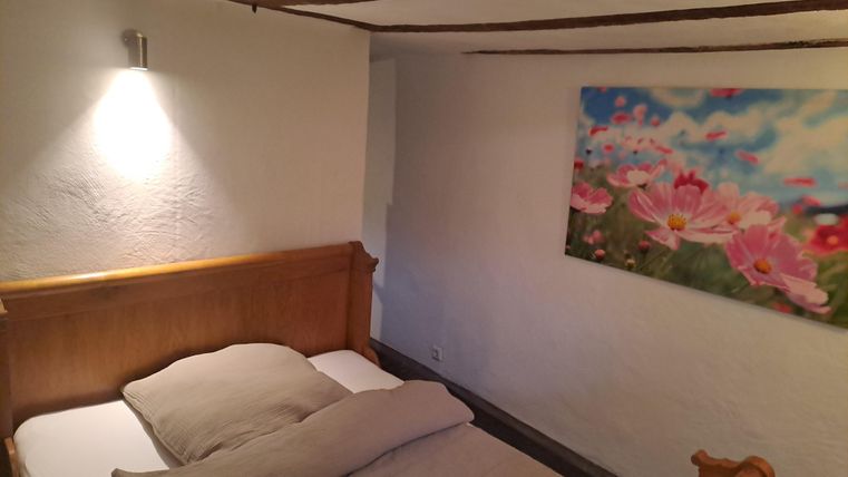 Ein gemütliches Schlafzimmer mit einem Holzbett und neutraler Bettwäsche. An der Wand hängt ein buntes Bild mit Blumenmotiven.