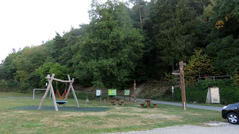 Ein Spielplatz mit einer Schaukel und einigen Sitzgelegenheiten in einem grünen Bereich. Im Hintergrund sind Bäume und ein Weg zu sehen.