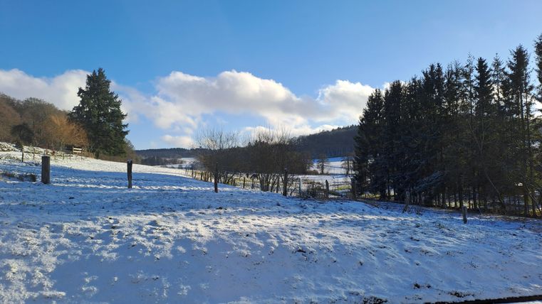 Eine verschneite Landschaft unter einem klaren blauen Himmel. Im Hintergrund sind Bäume und sanfte Hügel zu sehen.
