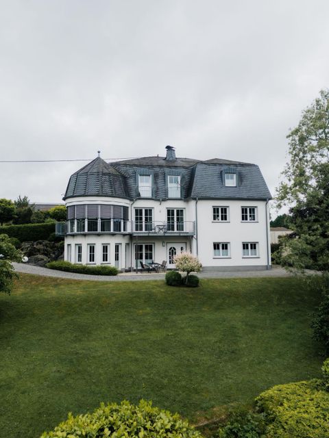 Ein großes, elegantes Haus mit einem grauen Dach und weißen Wänden. Der Garten ist gepflegt und grün, und der Himmel ist bewölkt.