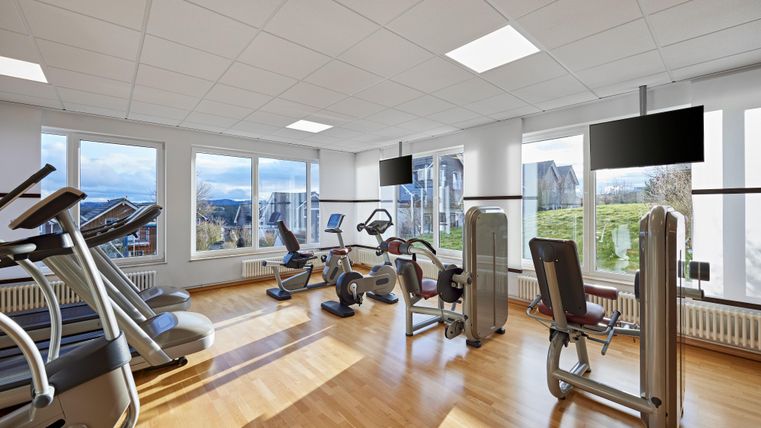 Ein modernes Fitnessstudio mit verschiedenen Trainingsgeräten. Große Fenster sorgen für viel Tageslicht und einen Blick ins Freie.