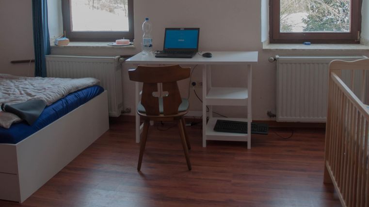 Ein einfaches Zimmer mit einem Bett, einem Schreibtisch und einem Kinderbett. Der Raum hat große Fenster, die natürliches Licht hereinlassen.