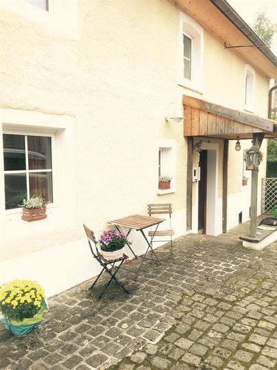 Ein gemütlicher Eingang zu einem Haus mit einem kleinen Tisch und Stühlen. Blumen in Töpfen verzieren die Fensterbänke und den Eingangsbereich.