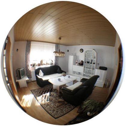 Ein gemütliches Wohnzimmer mit schwarzen Sofas und einem weißen Tisch. Es gibt Fenster mit Gardinen und einen Fernseher in der Ecke.