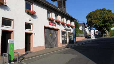 Eine ruhige Straße mit einem weißen Gebäude und blühenden Blumenkästen. Im Vordergrund steht eine Ladesäule für Elektroautos.