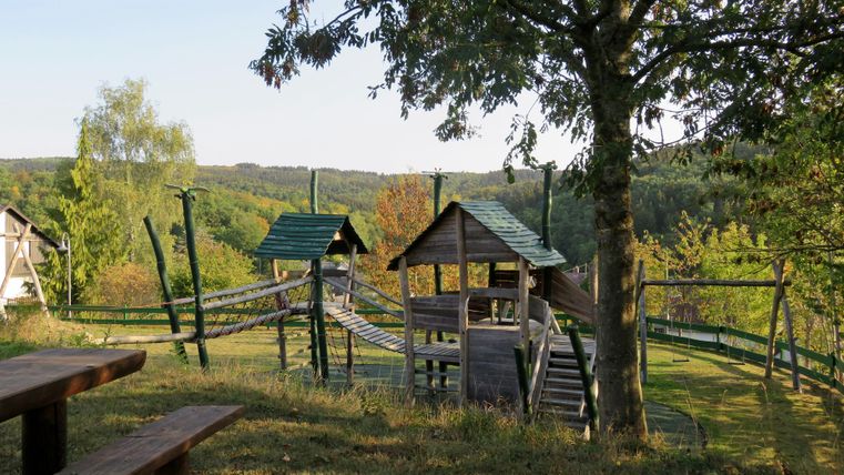 Ein Spielplatz mit Holzgerüsten und Rutschen in einer grünen Landschaft. Im Hintergrund sind Bäume und Hügel sichtbar.