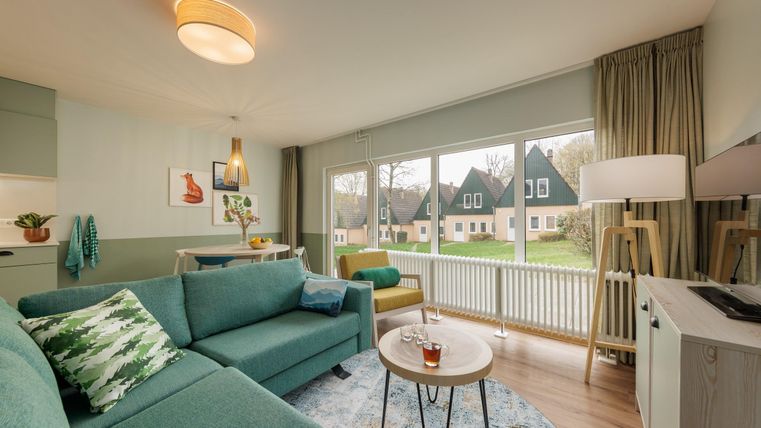 Eine moderne Wohnzimmereinrichtung mit einem gemütlichen Sofa und einem runden Tisch. Große Fenster bieten einen Blick auf einen grünen Garten und Häuser im Hintergrund.