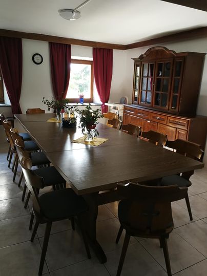 Ein großer Essbereich mit einem langen Holztisch und vielen Stühlen. Der Raum hat Fenster mit roten Vorhängen und eine Vitrine im Hintergrund.