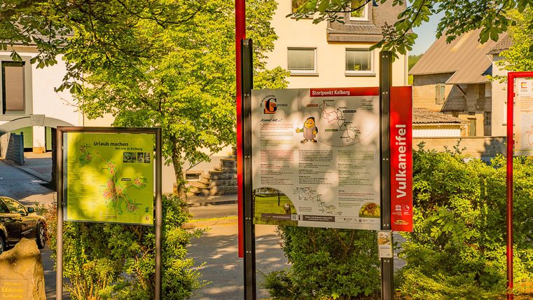 Ein Informationstafel steht zwischen Bäumen und Häusern. Die Umgebung wirkt grün und einladend.