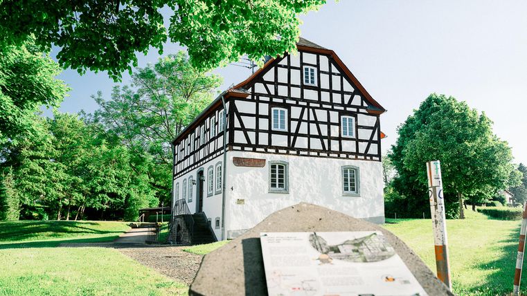 Ein historisches Fachwerkhaus umgeben von Bäumen und Grünflächen. Im Vordergrund ist ein Informationsschild zu sehen.